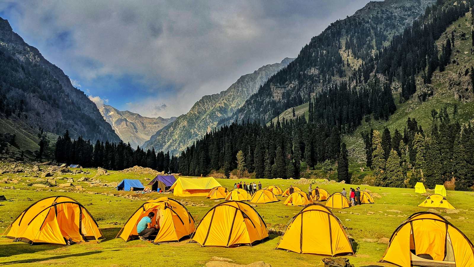 Rows of yellow tents pitched in a green mountain valley under a cloudy sky